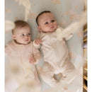 Two babies in soft, sky-themed sleep & play outfits lie on a cloud-patterned crib mattress, one gazing curiously.