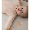 Baby wearing a pink sleep and play outfit on a patterned surface with clouds and moons, looking curiously at the camera.