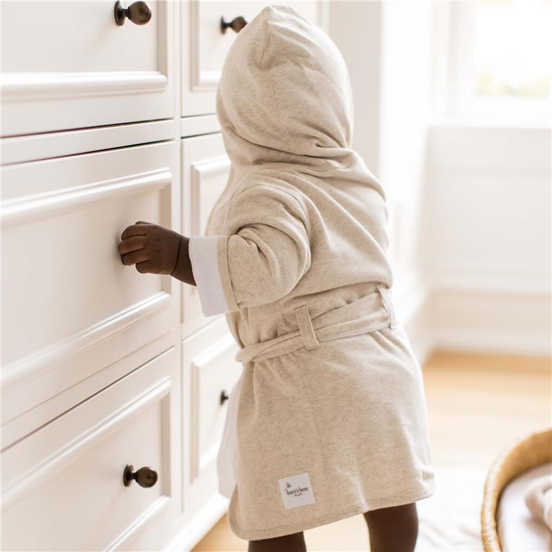 Hooded oatmeal robe for babies, featuring a belt and white cuffs, as a child explores a white dresser.