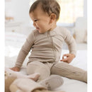 Baby wearing a beige ribbed one-piece outfit, smiling while sitting on a bed with a stuffed animal nearby.