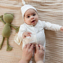 Baby wearing a Burts Bees Frog Jumpsuit and hat, with a green frog plush toy nearby, as a hand adjusts the outfit.