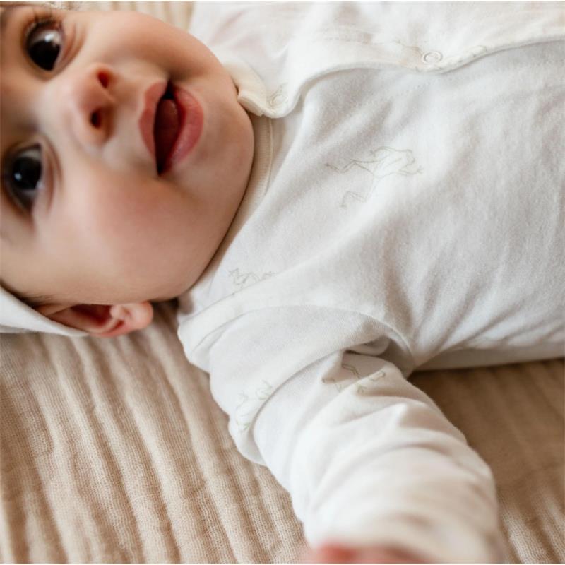 Baby wearing a Burts Bees white jumpsuit with subtle frog prints and a matching hat, smiling on a soft, textured surface.