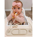 A baby in a high chair holds a yellow teething toy shaped like a banana, wearing a blush pink headband and shirt.