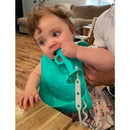 A baby in a high chair bites a bright spearmint Busy Baby Bungee Bib while being held by an adult.