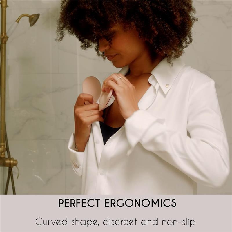 A woman in a white shirt is inserting a beige nursing pad into her top, showcasing its ergonomic, discreet, and non-slip design.