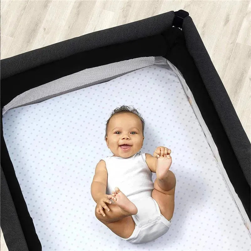 Baby lying on a pink dotted playard sheet with a smiling expression, inside a black playard.