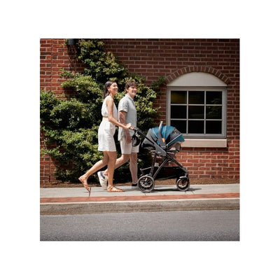 A couple walks on a sidewalk with the Chicco Corso Primo Cleartex Modular Travel System in Hazelnut, featuring a stylish stroller with a blue canopy.