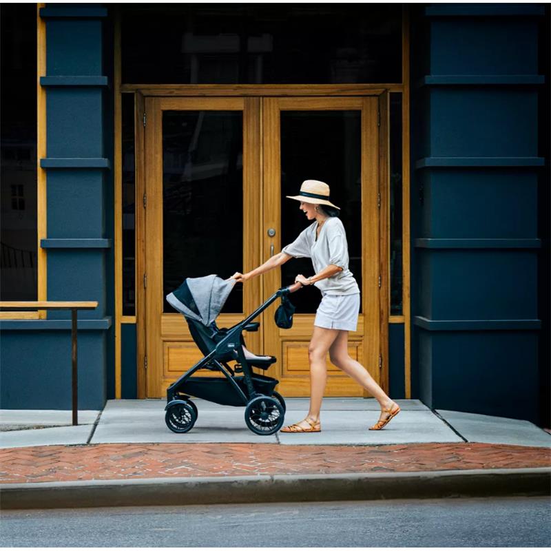 A woman in a hat walks down the sidewalk, pushing a Chicco Corso Primo ClearTex stroller with a gray canopy.