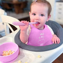 Chicco Easy Start Silicone Feeding Gift Set in pink, featuring a baby in a high chair holding a spoon and sitting beside a bowl of snacks.