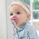 Toddler with a pink Chicco Physioforma Dual Soft Pacifier attached to a decorative strap, wearing a light blue shirt and headband.