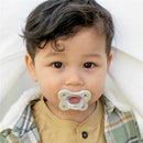 Child with curly hair holding a grey and dusty rose pacifier, wearing a beige shirt and checkered jacket.
