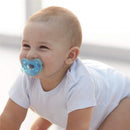 Smiling baby crawling while wearing a white onesie and using a blue/green silicone pacifier.