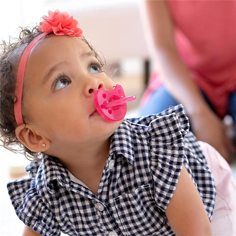 A toddler with curly hair and a pink headband looks up while holding a dark pink orthodontic pacifier in her mouth.