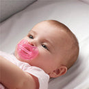 Baby with a pink orthodontic silicone pacifier, lying on a soft surface, wearing a light pink shirt.