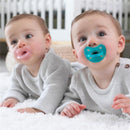 Two infants wearing light gray sweaters, one with a soft pink pacifier and the other with a turquoise pacifier, on a white fluffy surface.