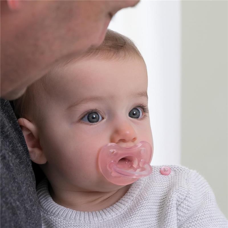 Baby with light pink orthodontic silicone pacifier, wearing a white sweater, looking curiously with blue eyes.