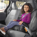 Backless booster car seat by Chicco with a girl sitting in it, wearing a purple shirt and floral leggings, securely buckled in a vehicle.