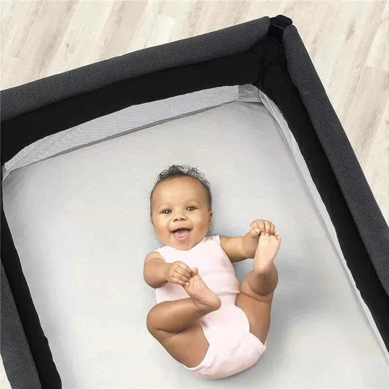 Baby lying on a Chicco Lullaby Playard Sheet in Ivory, smiling and playing with feet in a playard setting.