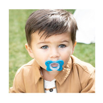 Child with light brown hair and blue eyes holds a blue Chicco Physioforma pacifier in a park setting.