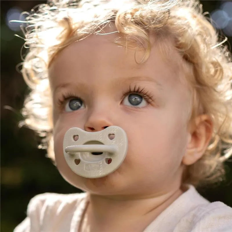 Chicco Physioforma Luxe pacifier in grey held by a baby with curly hair, looking up with blue eyes.