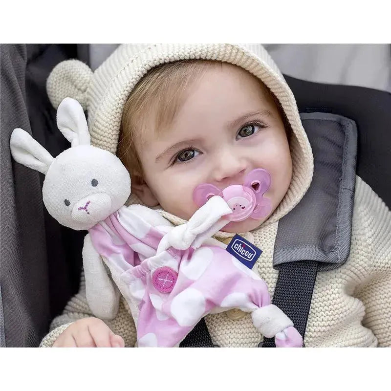 Toddler wearing a cream hoodie, holding a Chicco Pocket Buddy toy with a pink pacifier in mouth and smiling.