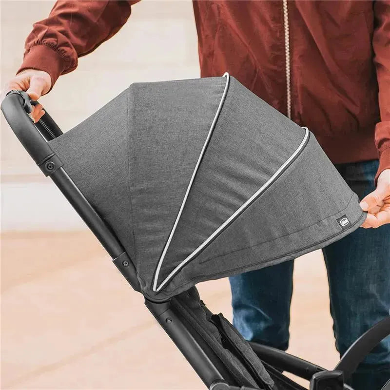 Self-folding compact stroller in graphite, being adjusted by a person. Features a large canopy and sturdy frame.