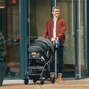 Man smiling while pushing a Chicco Presto Self-Folding Compact Stroller in graphite color, exiting a commercial building.