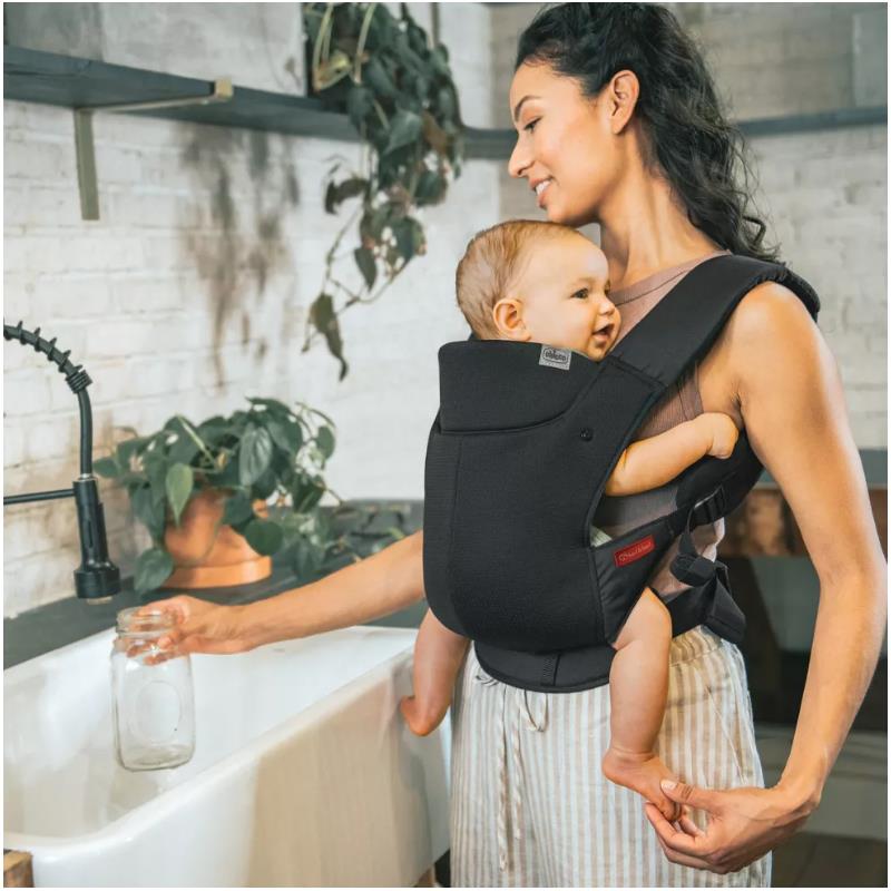 Chicco Snugsupport 4-In-1 Infant Carrier in black, worn by a woman holding a jar near a sink, with a baby comfortably seated inside.
