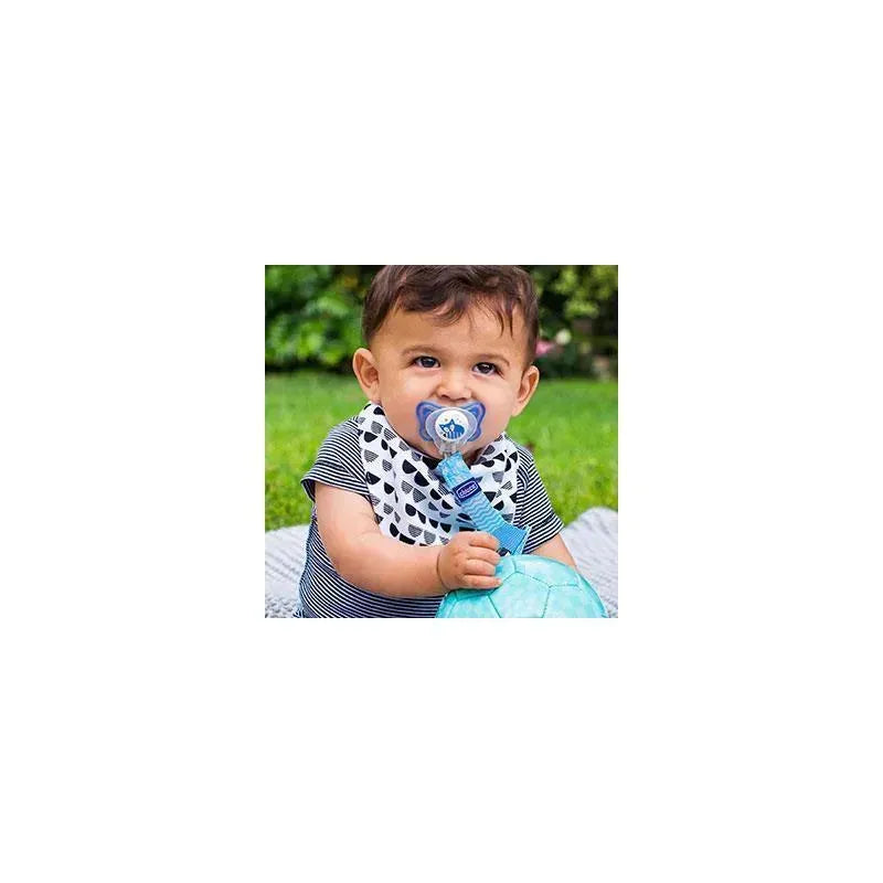 Baby sitting on grass, holding a teal ball, wearing a striped shirt with a pacifier attached to a grey Chicco clip.