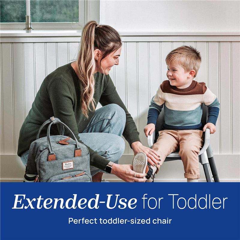 A woman helps a young boy put on shoes while he sits in a toddler chair, with a diaper bag nearby.