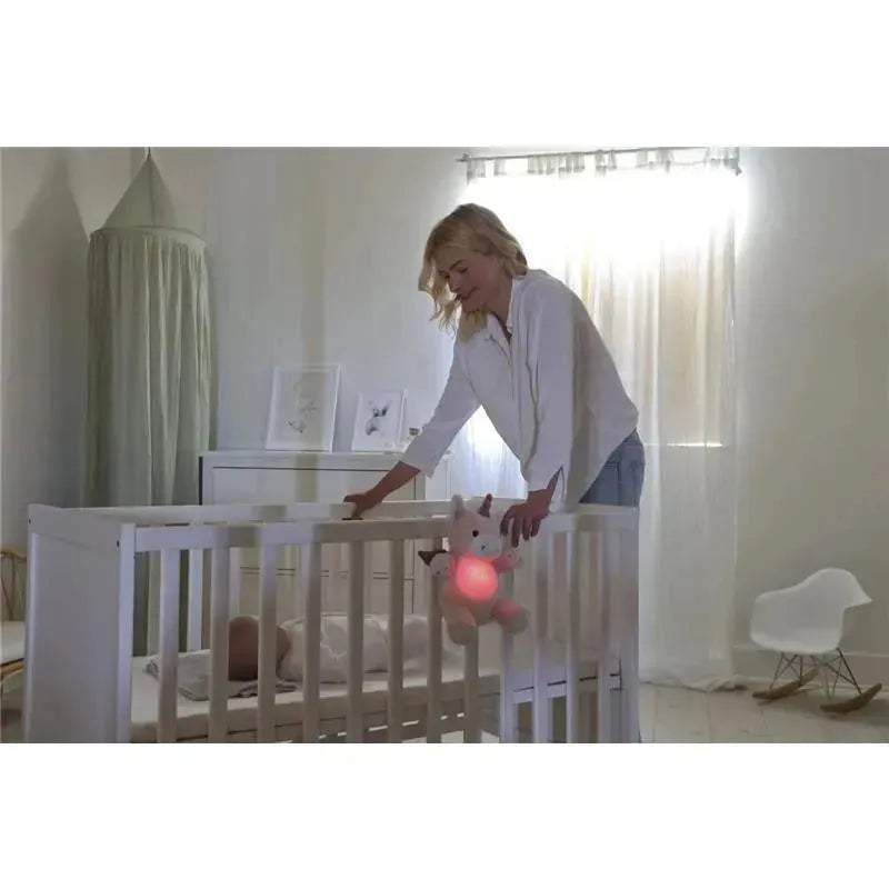 A woman gently reaches into a crib where a baby sleeps, with a unicorn night light attached to the crib, glowing softly pink.
