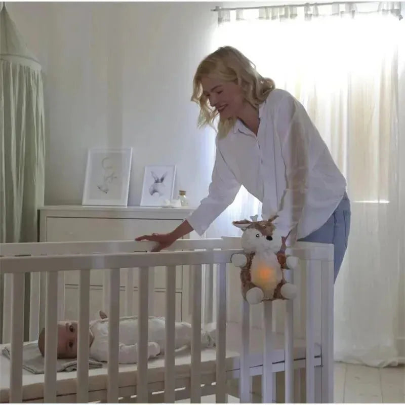 A woman stands beside a crib, smiling at a baby lying inside, with a Cloud B Love Light Buddy, Finley Fawn, glowing softly nearby.