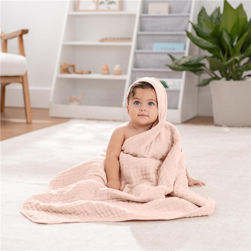Hooded baby towel in mauve and blush draped around a sitting baby with blue eyes, in a cozy room with plants.