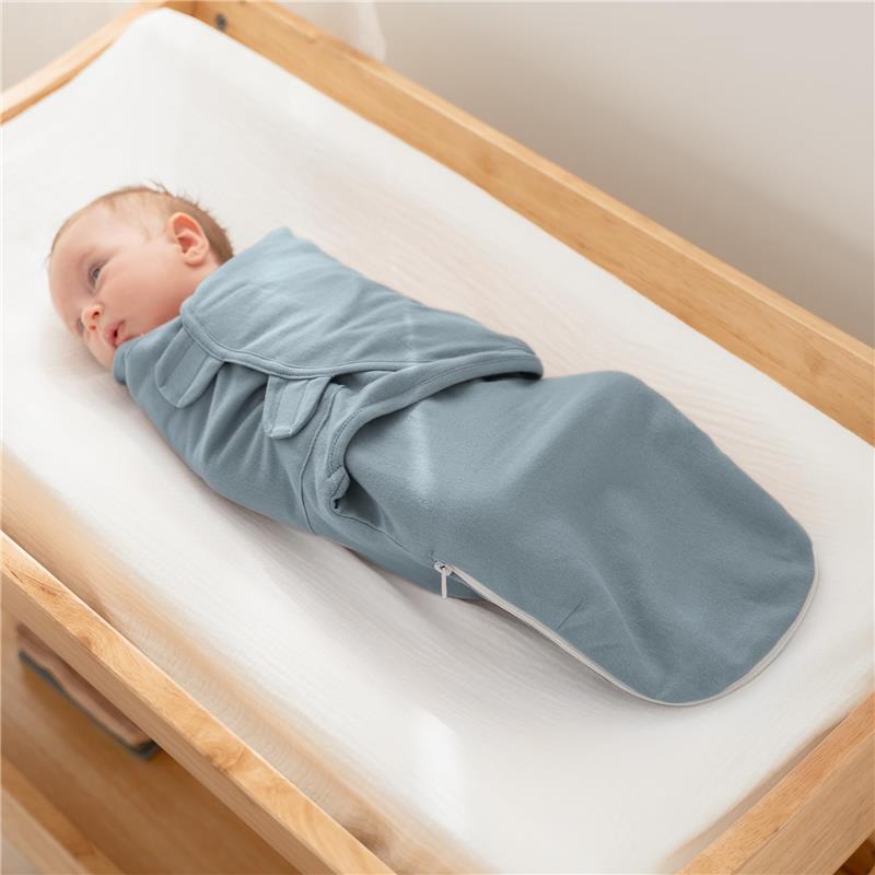 Baby swaddled in a soft blue blanket with zipper closure, lying on a crib mattress in a wooden crib.