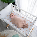 Baby swaddled in a soft, blush muslin blanket lying in a crib with a rainbow-printed mattress pad.