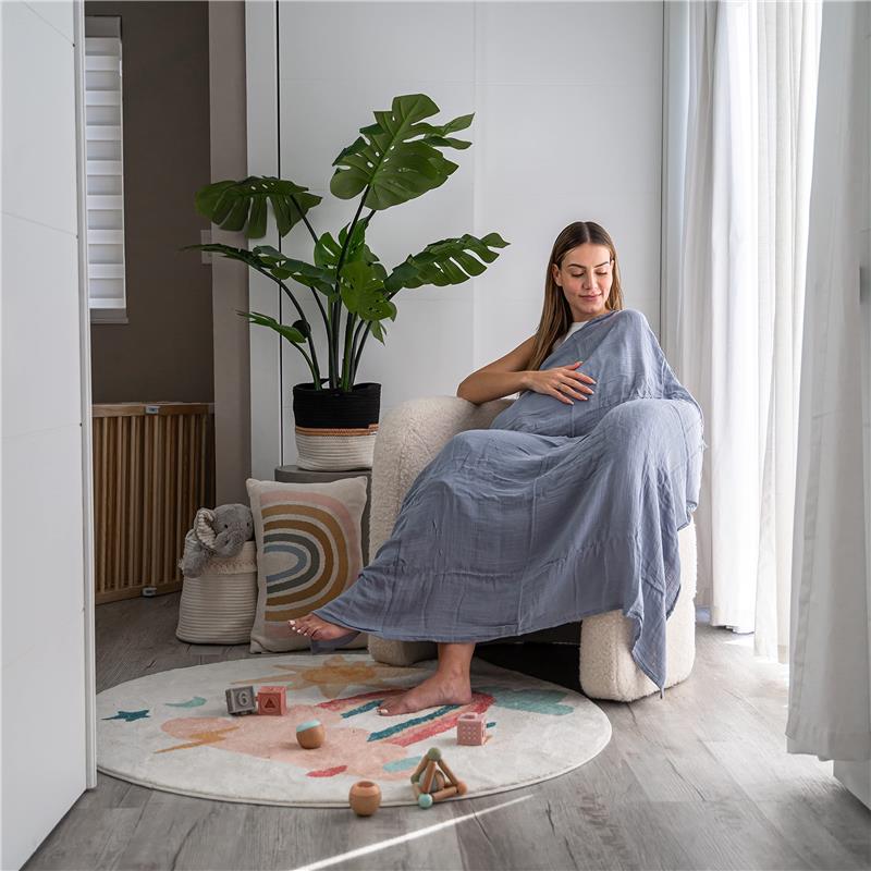 A woman draped in a blue muslin swaddle blanket sits on a chair, surrounded by indoor plants and colorful toys on a rug.