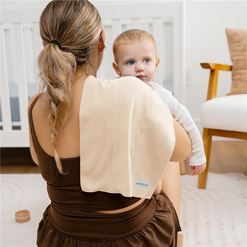 A woman holds a baby while a cream-colored Comfy Cubs burp cloth rests on her shoulder, with a nursery setting in the background.