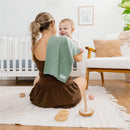 Mom holding a smiling baby, using a green burp cloth, in a warmly decorated nursery with toys and a crib.
