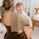 A woman holds a child on her shoulder, with a beige burp cloth draped over her shoulder, in a cozy, bright room.
