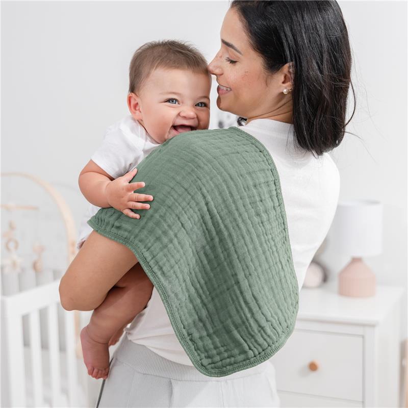 Mom holds a smiling baby over her shoulder, using a soft green muslin burp cloth. The background features a nursery.