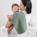Mom holds a smiling baby over her shoulder, using a soft green muslin burp cloth. The background features a nursery.