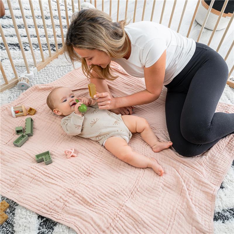A mother plays with her baby on a blush Comfy Cubs muslin blanket, surrounded by colorful toys.