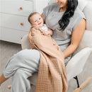 A smiling baby wrapped in a cream Comfy Cubs muslin blanket, held by a woman in a cozy chair.