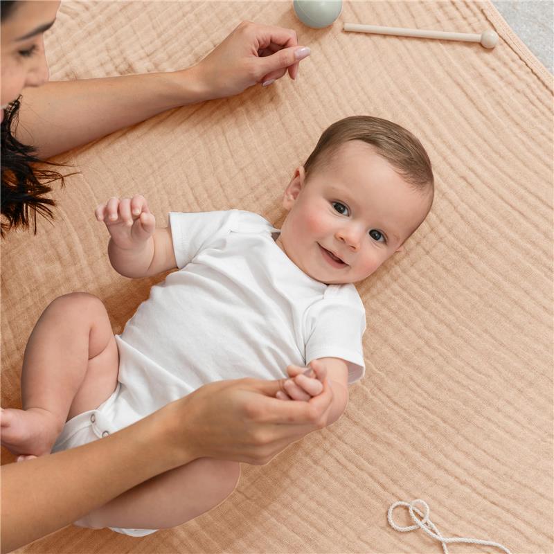 Baby in a white onesie smiles while being played with by an adult, on a cream-colored muslin blanket.