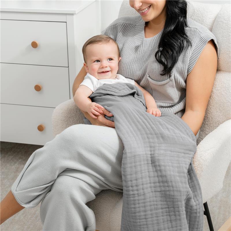 A smiling baby wrapped in a grey Comfy Cubs Muslin Baby Blanket, held by a woman in comfortable clothing, sitting in a cozy chair.