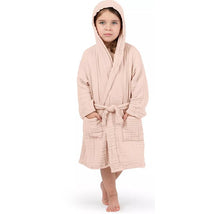 Muslin kids bathrobe in blush color, featuring a hood and pockets, worn by a girl standing barefoot.