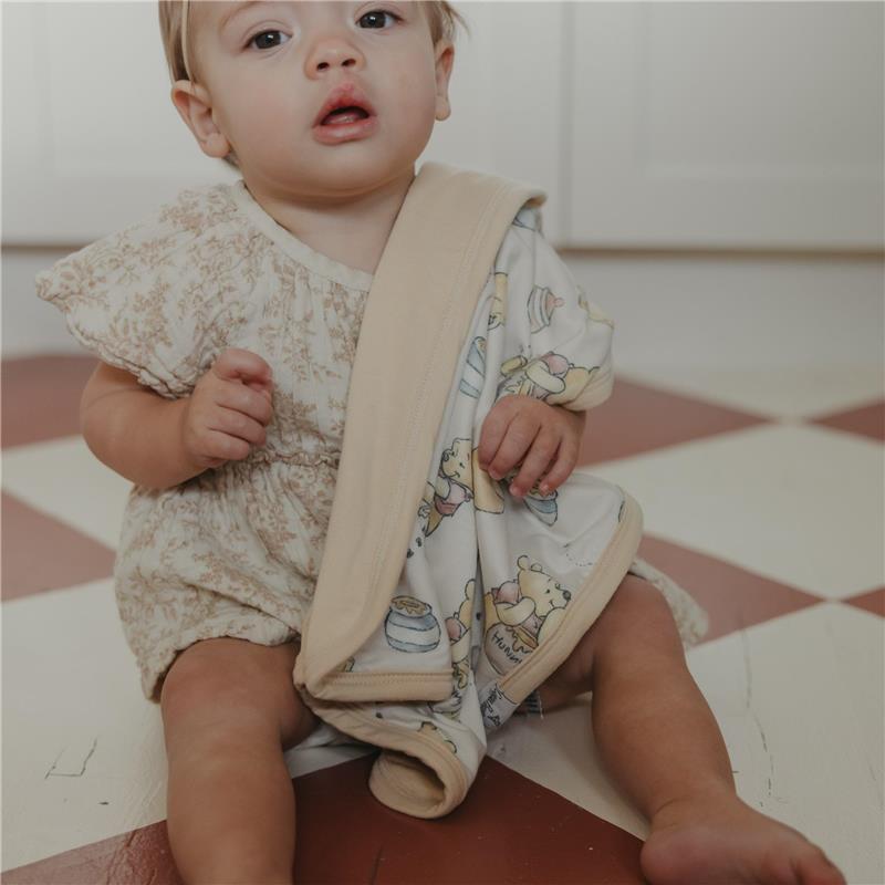 Toddler in a floral outfit holding a soft security blanket featuring a Winnie the Pooh design and beige trim.