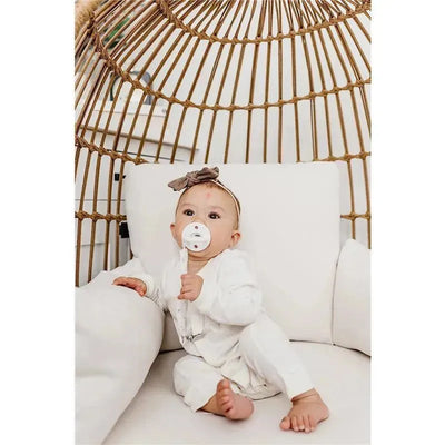 Baby sitting in a white chair, wearing a white outfit and a bow headband, holding a pacifier clip.