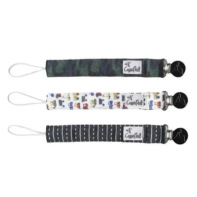 Three baby binky clips from Copper Pearl in different patterns: camo, vehicle print, and road design, featuring metal clips.