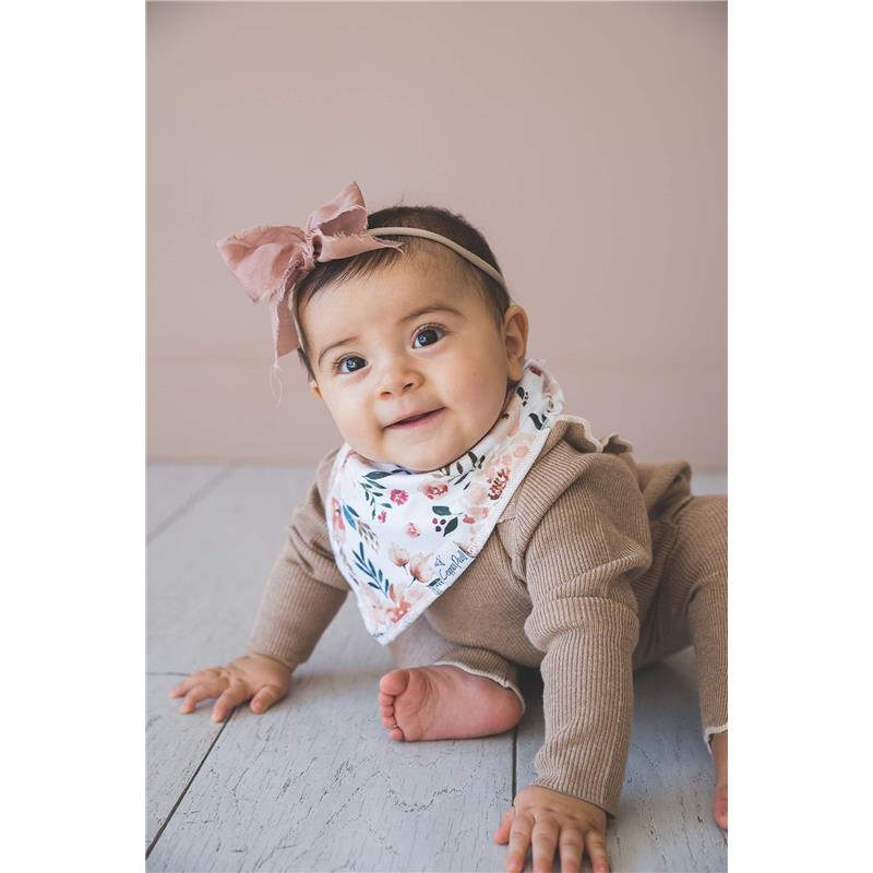 Baby wearing a floral bandana bib sits on hardwood floor, smiling while sporting a pink bow headband.